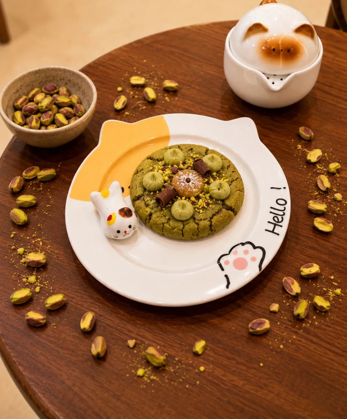 Un cookie à la pistache sur une assiette blanche en forme de tête de chat, avec Hello! écrit sur le bord et un petit chat en céramique à côté.