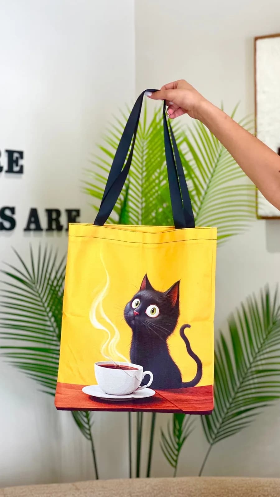 Une main tenant un tote bag jaune imprimé d'un chat noir attablé, devant le mur du café portant COME WHERE #CATS ARE.